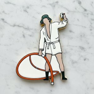May include: A cookie shaped like a figure wearing a white bathrobe, a green hat, and black boots, holding a small black device in one hand and a large orange and white circular object in the other, with black outlines defining the details.