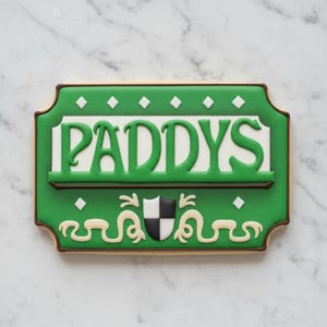 May include: A rectangular cookie with green icing and the word "PADDYS" in green. The cookie has a white background for the text, a brown border, and decorative elements, including a shield.