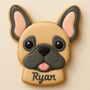 May include: A tan-colored cookie shaped like a French Bulldog's face, with black eyes, nose, and accents. The dog's ears are outlined in gray with pink inner ears. The name "Ryan" is written in cursive at the bottom.
