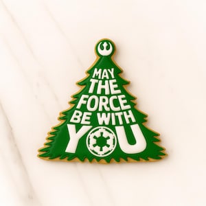 May include: A Christmas tree-shaped biscuit decorated with green icing and the words "MAY THE FORCE BE WITH YOU" in white. The biscuit also features the Rebel Alliance and Galactic Empire symbols. A festive treat for Star Wars fans.