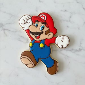 May include: A decorated sugar cookie shaped like the video game character Mario. The cookie has a red hat with a white "M", blue overalls, a brown moustache, and white gloves. The cookie is on a white marble surface.