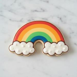 Puede incluir: Una galleta con forma de arcoíris con un diseño colorido. La galleta presenta un arcoíris rojo, naranja, amarillo, verde y azul, con acentos blancos en forma de nube delineados en marrón. La galleta está sobre un fondo de mármol blanco.