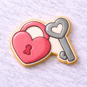 Op de afbeelding: Een hartvormige koekje versierd met roze glazuur, dat lijkt op een hangslot, naast een grijze sleutel met een hartvormige kop. De koekje heeft een gele omtrek en ligt op een wit marmeren oppervlak. De sleutel is gepositioneerd om het hartvormige hangslot te ontgrendelen.