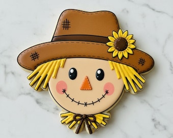 May include: A scarecrow-shaped sugar biscuit decorated with royal icing. The design features a brown hat with a yellow sunflower, yellow straw hair, a triangular orange nose, pink cheeks, and a stitched smile with black thread details.