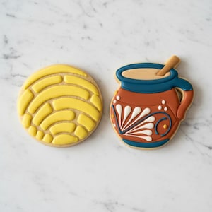 May include: Two decorated sugar biscuits. One is a round yellow biscuit with a textured, shell-like pattern. The other is a brown mug-shaped biscuit with a blue rim, a tan top, and white decorative floral-inspired icing details on the front.
