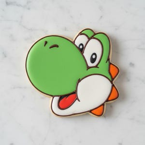 Peut inclure: Un biscuit vert en forme de Yoshi, avec des détails blancs, oranges et rouges. Le biscuit a un contour marron et est décoré de glaçage. Sur une surface en marbre blanc.