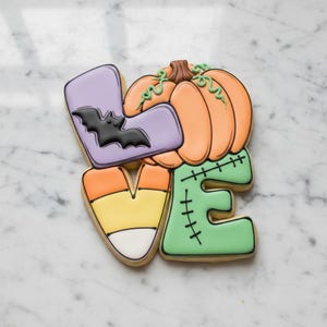 May include: A decorative cookie shaped like the word LOVE, featuring a light purple L with a black bat, an orange pumpkin O, a candy corn V, and a light green E with black stitched detailing, all decorated with smooth royal icing.