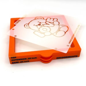 Op de afbeelding: Een oranje koekjessnijderframe met een doorzichtige plastic sjabloon met een berenontwerp. De sjabloon heeft het woord "LOVE" en de website "EZCOOKIECUTS.COM" erop gedrukt. Het frame is gelabeld "TOP EXTENSION 0,64 cm BASE 0,64 cm".