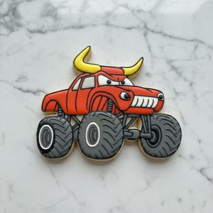 May include: A decorated sugar cookie in the shape of a red monster truck with a bull's head. The truck has large gray tires with white accents and yellow horns. The cookie is on a white and gray marble surface.
