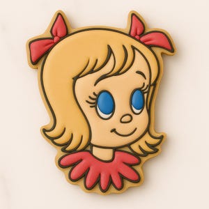 May include: A decorated cookie in the shape of a cartoon girl's face. The cookie features blonde hair, blue eyes, and a red bow. The girl is wearing a red dress. The cookie is outlined in black and has a light brown border.