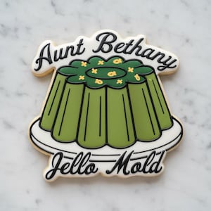 Può includere: Un biscotto a forma di stampo per gelatina verde scanalato su un piatto bianco. Il testo Aunt Bethany è scritto in glassa nera nella parte superiore, e Jello Mold è scritto in glassa nera nella parte inferiore. Lo stampo verde ha piccoli accenti gialli.