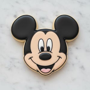 以下が含まれることがあります： ミッキーマウスの形をしたクッキー。黒い耳、黒い鼻、笑顔の顔があります。クッキーは薄いタン色の顔と黒い輪郭をしています。クッキーは白い大理石の表面にあります。