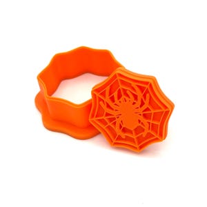 May include: Bright orange Halloween-themed cookie cutter set. The set includes a round cutter and a spider web-shaped stamp with a spider in the center. Perfect for making festive cookies for the spooky season.