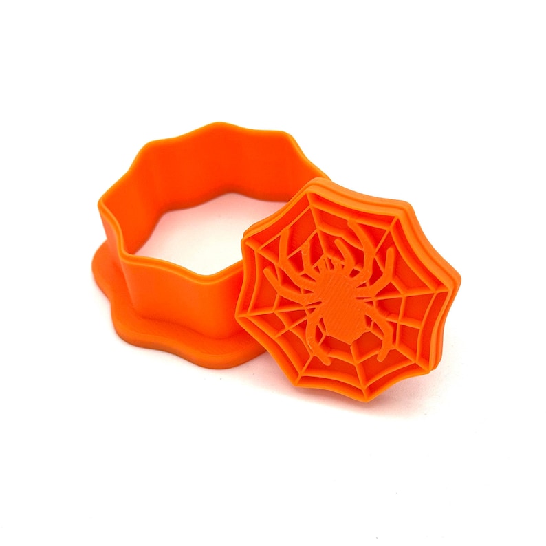 May include: Bright orange Halloween-themed cookie cutter set. The set includes a round cutter and a spider web-shaped stamp with a spider in the center. Perfect for making festive cookies for the spooky season.