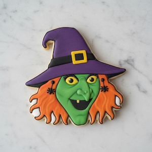 Puede incluir: Una galleta con forma de cara de bruja, con cabello naranja brillante, tez verde y un sombrero puntiagudo morado con una banda negra y una hebilla amarilla. La bruja tiene pendientes en forma de araña y un solo diente amarillo visible.