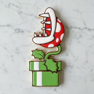 Könnte beinhalten: Ein dekorierter Zuckerkeks in Form einer Piranha-Pflanze aus der Super Mario-Videospielserie. Der Keks hat einen roten Kopf mit weißen Punkten, weiße Zähne und einen grünen Stiel und eine grüne Box. Die Box hat weiße Streifen.