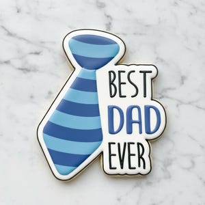 Puede incluir: Una galleta de azúcar decorada con forma de corbata, con rayas azules y celeste. Las palabras "BEST DAD EVER" están escritas en glaseado negro y azul.
