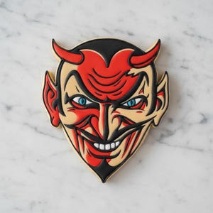 May include: A cookie shaped like a devil face with red, black, and cream-colored icing. The design features pointed ears, horns, a sharp chin, and blue eyes with a wide, toothy grin outlined in bold black lines on a smooth surface.