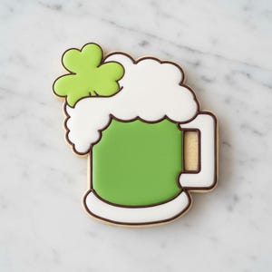 May include: A St. Patrick's Day cookie shaped like a green beer mug with white foam and a green shamrock. The cookie has a brown outline and is set against a white marble background.