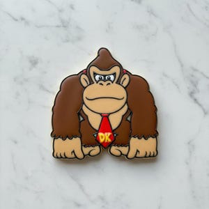 Peut inclure: Image de Donkey Kong, un personnage de gorille de style dessin animé. Le gorille est brun avec des mains et des pieds beiges, portant une cravate rouge avec "DK" en jaune. L'image a un contour blanc et est posée sur un fond de marbre blanc.