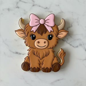 May include: A decorated cookie in the shape of a Highland cow. The cookie is brown with a pink bow on its head. The cow has large black eyes and a smiling face. The cookie is sitting on a white marble surface.