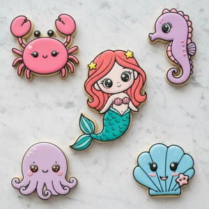 May include: A collection of decorated sugar cookies in a marine theme. The cookies include a pink crab, a mermaid with red hair and a teal tail, a purple seahorse, a purple octopus, and a blue seashell with a starfish.