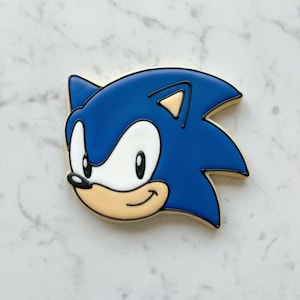 Può includere: Un biscotto blu, bianco e beige a forma di testa di Sonic the Hedgehog. Il biscotto presenta un design dettagliato con punte blu, occhi bianchi e un muso beige. Il biscotto è bordato di nero.