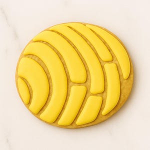 May include: A round, golden-brown cookie with a yellow, shell-like design on top. The yellow design is made of smooth, curved shapes, creating a textured appearance. The cookie is set against a white marble surface.