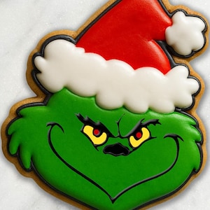 May include: A sugar cookie shaped like a green character face with a frowning expression, yellow eyes with red pupils, and black outlines. The character wears a red Santa hat with white icing trim and a white pom-pom, all decorated with royal icing.