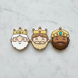 May include: Three decorated sugar cookies shaped like kings' heads, each wearing a yellow crown with a small gem. The first has a white beard, the second a light brown beard, and the third a dark brown beard, all with smiling expressions.