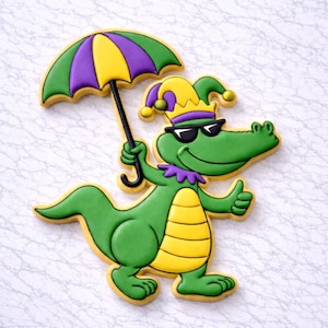 Könnte beinhalten: Ein dekorierter Keks in Form eines grünen Alligators mit Sonnenbrille, Narrenhut und lila Kragen, der einen gelben, grünen und lila Regenschirm hält. Der Alligator gibt einen Daumen hoch.