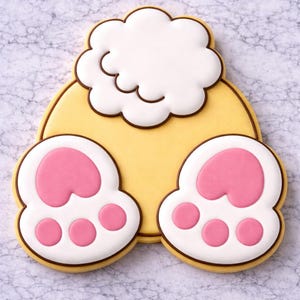 May include: A whimsical cookie shaped like a bunny's rear, featuring a yellow body, white fluffy tail, and two white paws with pink pads and brown outlines. The cookie is decorated with icing.