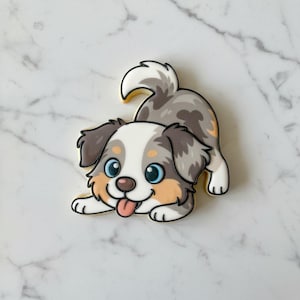 May include: A sugar cookie shaped like a playful puppy with a merle coat pattern in shades of gray, white, and tan. The puppy has large blue eyes, a dark brown nose, and a small pink tongue sticking out, outlined in black icing.
