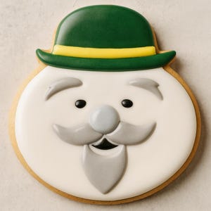 Könnte beinhalten: Ein St. Patrick's Day-Keks in Form eines Kobolds. Der Keks hat einen grünen Hut mit gelbem Band, ein weißes Gesicht mit grauem Schnurrbart und Bart sowie schwarze Augen. Der Keks ist goldbraun umrandet.