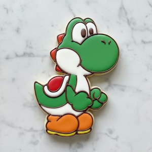 May include: A decorated cookie shaped like Yoshi, the green dinosaur character from the Super Mario video game series. The cookie has green, white, red, orange, and brown icing with a yellow outline. The cookie is standing upright.
