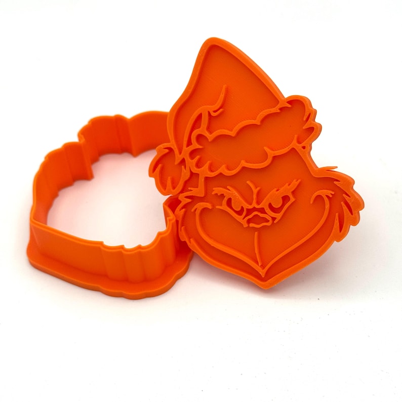 May include: An orange plastic cookie cutter set featuring a Grinch face wearing a Santa hat. The set includes the Grinch's face and a heart-shaped outline. The cookie cutters are made of orange plastic.