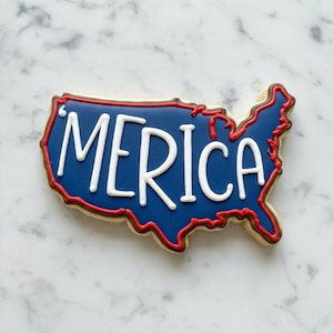 May include: A cookie shaped like the United States, with a blue interior and red outline. The word "MERICA" is written in white across the center of the cookie. The cookie has a light brown border.