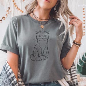 Peut inclure: T-shirt gris avec un dessin au trait noir d'un chat grincheux. Le chat est assis avec une expression sévère. Le t-shirt a des manches courtes et une coupe décontractée, adapté pour un usage quotidien.