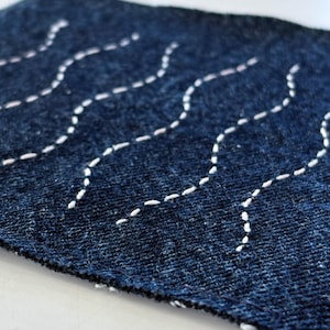 以下が含まれることがあります： 濃い青色の生地に、白い波状の手縫いの刺繍が施されたクローズアップ。ステッチは、質感のある素材の上に流れるようなパターンを作り出しています。生地の端が見え、その形状と構造を示しています。