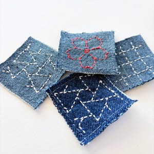 May include: Close-up of five square denim patches with white and red embroidery. The patches feature geometric and floral designs. The denim is a medium blue color, and the embroidery thread is white and red.