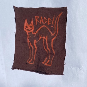 May include: A rectangular dark brown fabric patch with frayed edges featuring a screen-printed orange graphic of an arched-back cat with a skeletal face. The word RAGE! is printed in orange above the cat in a hand-drawn, jagged font style.