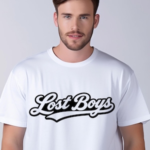 May include: White t-shirt with the text "Lost Boys" in a black, stylized font. The text has a white outline and a curved underline. The shirt is a crew neck style.