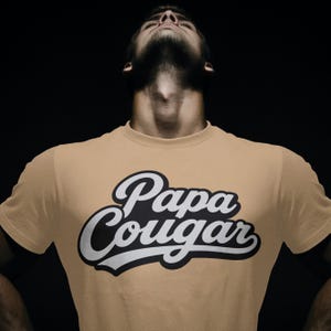 May include: Tan t-shirt with the words "Papa Cougar" in a black and white design. The shirt is made of a soft material and has short sleeves. The text is in a bold, stylized font.