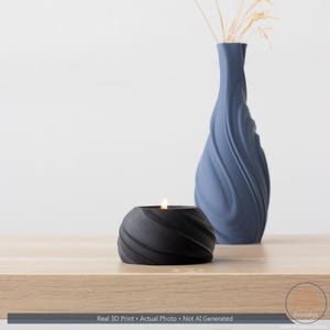May include: A black, spiral-textured candle holder with a lit candle is on a wooden surface. A blue vase with a similar spiral design and dried plants is in the background. The text at the bottom reads: Real 3D Print &bull; Actual Photo &bull; Not AI Generated.