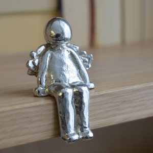 Pode incluir: Uma figura de prata de uma pessoa sentada com asas. A figura é feita de metal texturizado e tem um design simples.