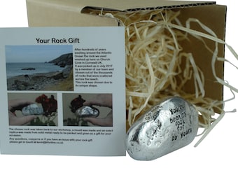 Puede incluir: Una roca de metal plateado con la inscripción "You've been my rock for 20 years" dentro de una caja de cartón con papel triturado. La roca es una réplica de una roca real que se encontró en una playa de Cornualles, Reino Unido.