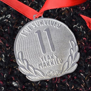 Op de afbeelding: Een zilveren medaille met de tekst "We Survived 11 Years Married" en een laurierkransontwerp.