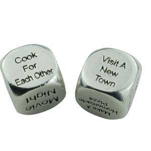 May include: Two silver dice with different phrases on each side. The first die has the phrases "Cook For Each Other" and "Movie Night". The second die has the phrases "Visit A New Town" and "Make A Homemade Pizza".