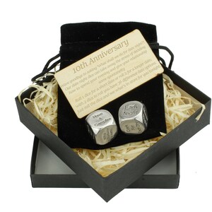 May include: A black gift box with a velvet bag inside. The bag contains two silver dice with engraved text. The text on the dice reads "Movie & Campfire" and "Early Night". A wooden card with the text "10th Anniversary" is visible in the bag. The card reads "Wave goodbye to asking 'What shall we do for date night?' Our date night dice can take away the stress of deciding how to spend your evening and open your relationship some spontaneity. Roll 1 dice for a short date or roll 2 for a longer date, with 2 dice to roll you have 36 different date options. Just roll the dice and see what's in store for you tonight."