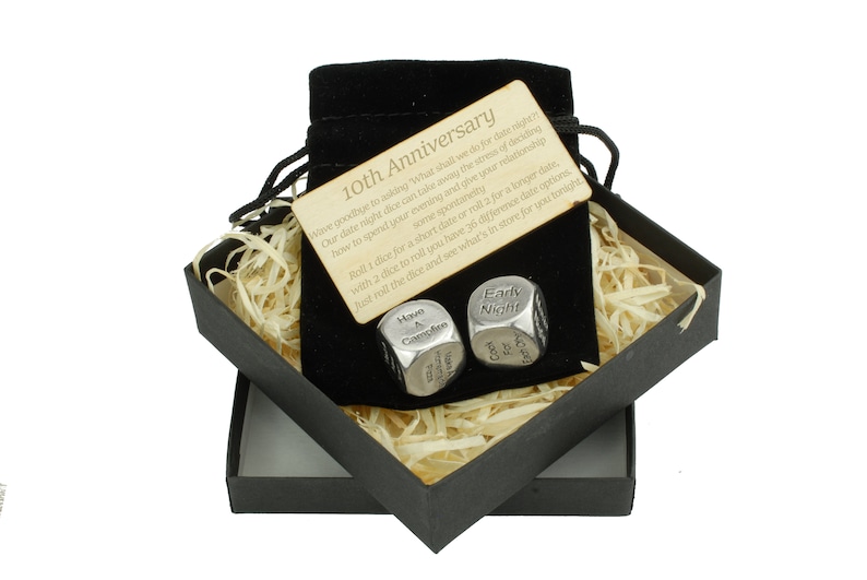 May include: A black gift box with a velvet bag inside. The bag contains two silver dice with engraved text. The text on the dice reads "Movie & Campfire" and "Early Night". A wooden card with the text "10th Anniversary" is visible in the bag. The card reads "Wave goodbye to asking 'What shall we do for date night?' Our date night dice can take away the stress of deciding how to spend your evening and open your relationship some spontaneity. Roll 1 dice for a short date or roll 2 for a longer date, with 2 dice to roll you have 36 different date options. Just roll the dice and see what's in store for you tonight."